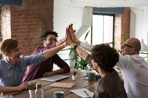 A group of colleagues sitting around a desk, all high-fiving each other at the same time, symbolizing teamwork and collaboration.