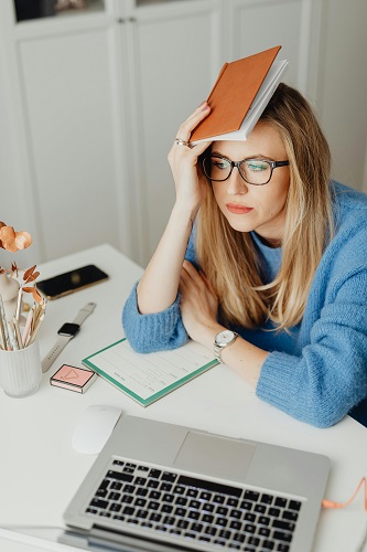 A stressed person balancing an open book on her head, sitting at a desk with an open notebook and laptop.